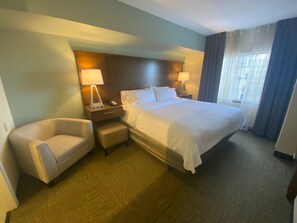 A hotel room with a large bed, a chair, a nightstand with a lamp, and a window with curtains.