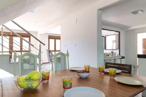 Un comedor con una mesa de madera, sillas, un cuenco de fruta y bebidas coloridas.