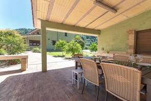 Um pátio externo com móveis de vime, uma mesa posta para refeições e vista para um edifício verde e árvores ao fundo.
