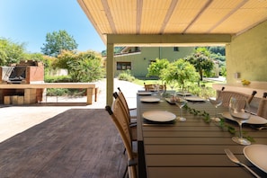 Un área de comedor al aire libre con una mesa preparada para cuatro personas, rodeada de sillas de mimbre, bajo un pérgola de madera.