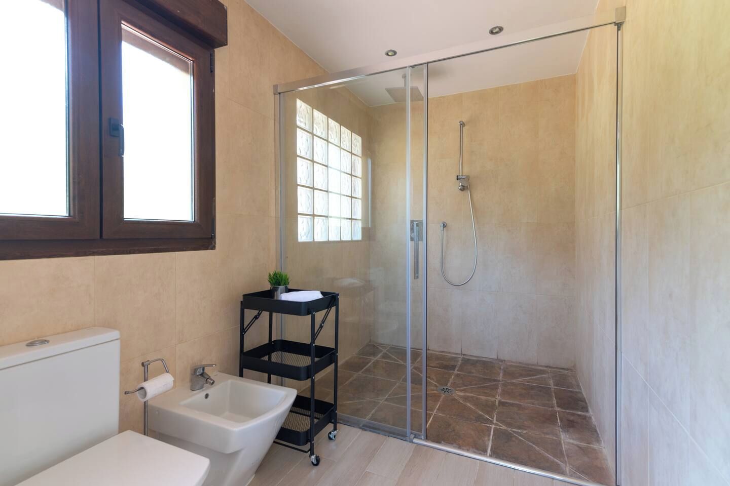 Een badkamer met een douchecabine van glas, een wit wastafelmeubel en een zwart plankje, en een raam.