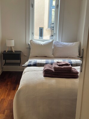 Een keurig opgemaakt bed met een witte dekbedovertrek en twee kussens, een klein nachtkastje met een lamp, en een raam met gordijnen.