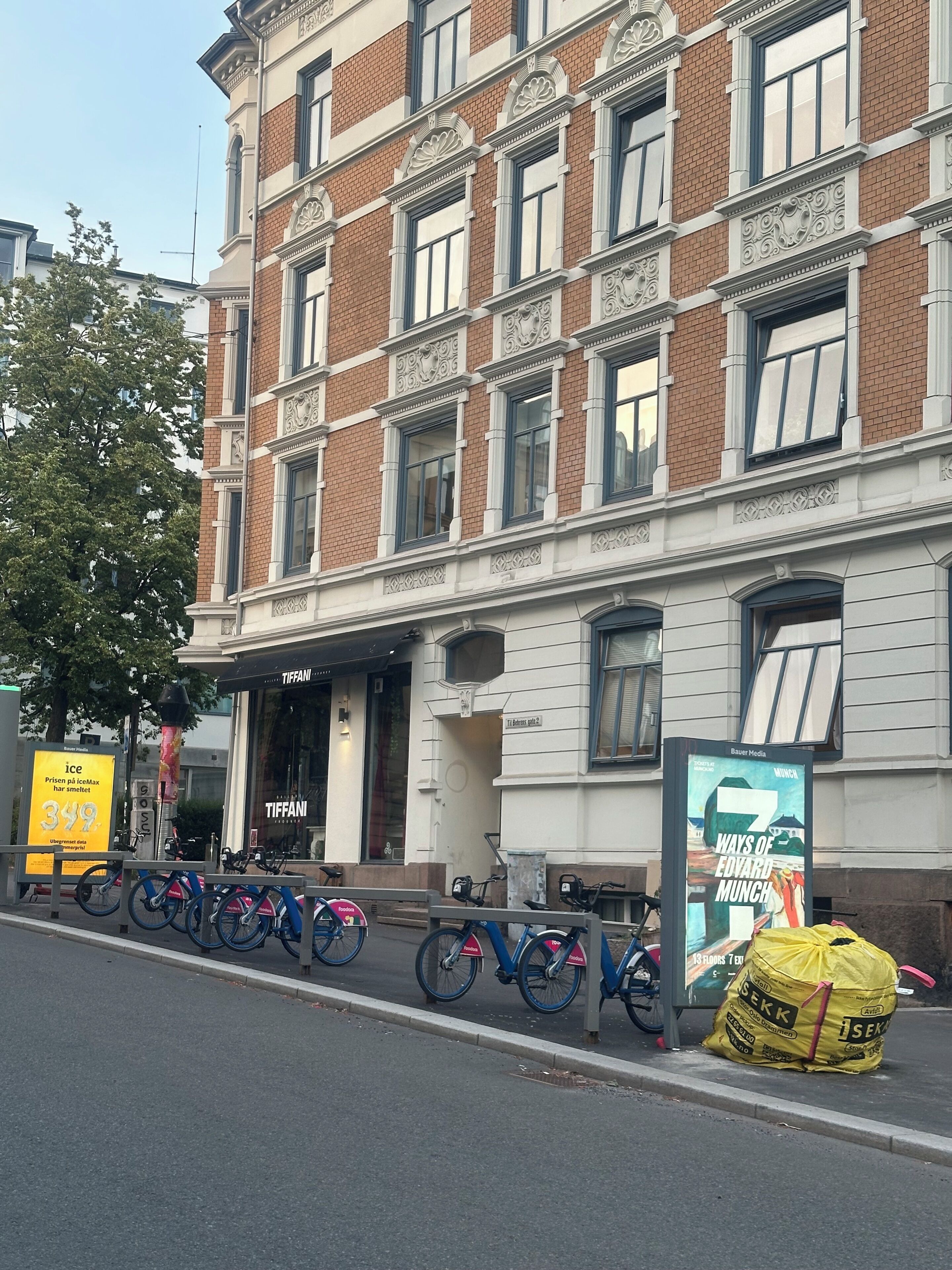 A building with a row of bicycles parked outside, a storefront with a sign, and a large advertisement on the sidewalk.