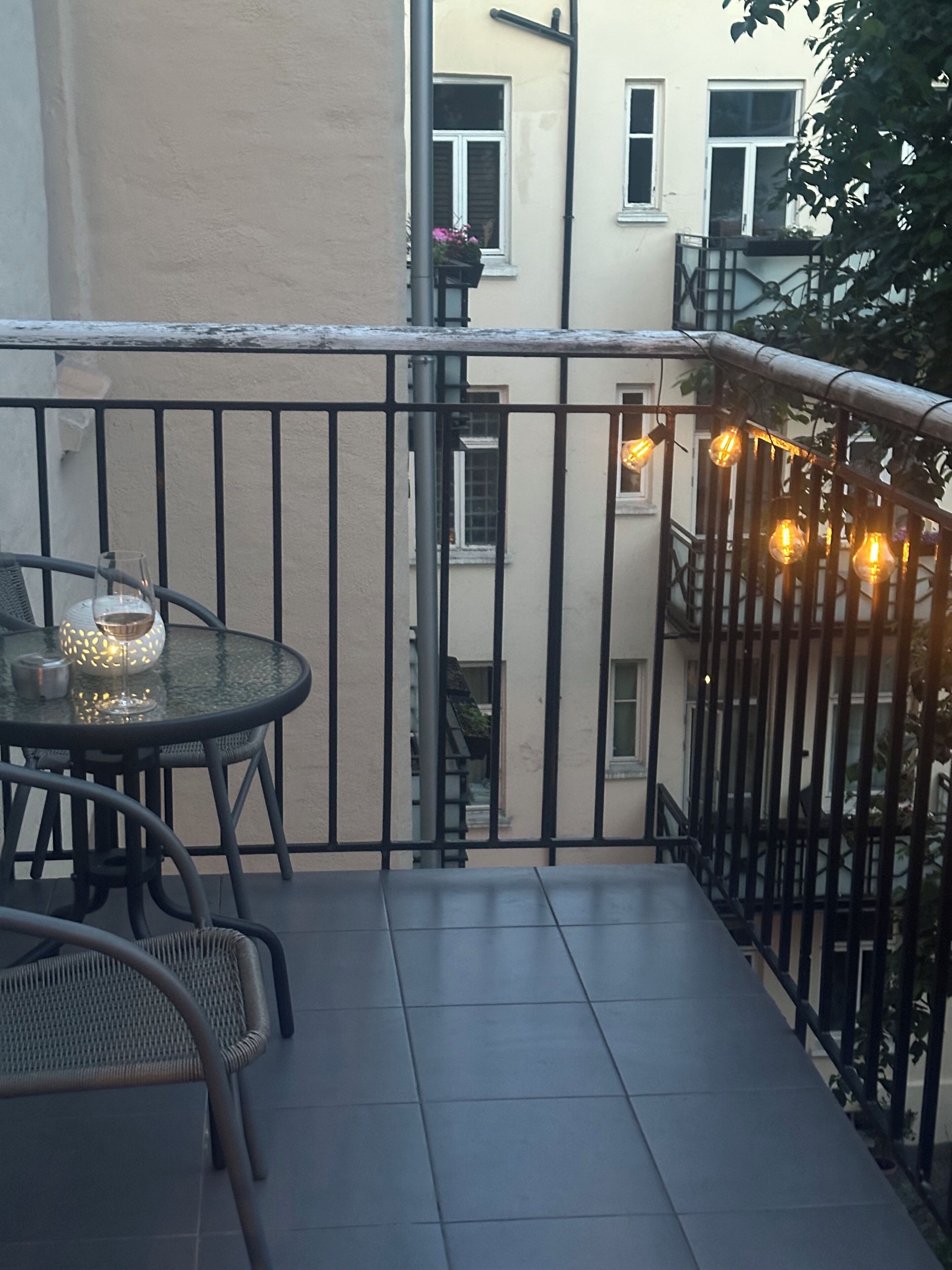 A balcony with a table and chairs, a hanging light fixture, and a view of adjacent buildings.