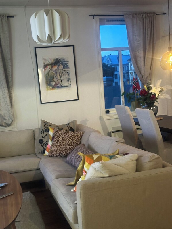 A living room with a sofa, a framed abstract painting, a window with curtains, and a vase with flowers.
