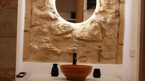 Une salle de bain avec un mur en pierre, un miroir rond, un lavabo blanc et un robinet noir.