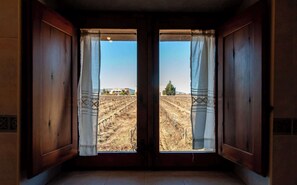 Une pièce aux murs en bois et dotée d’une grande fenêtre ornée de rideaux, qui offre une vue sur un champ aride et des bâtiments éloignés.
