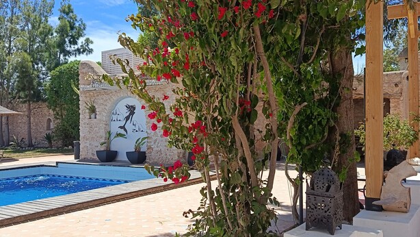 A poolside area with a stone wall, a wooden pergola, and a tiled table. - Villa 6 Chambres 3 Suites, piscine chauffée. Prix 18 pax avec petits déjeuners. (Essaouira)
