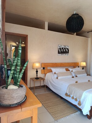 Un dormitorio con cabecera de madera, dos mesitas de noche con lámparas, una planta grande en maceta, una alfombra tejida y una obra de arte colgada en la pared.