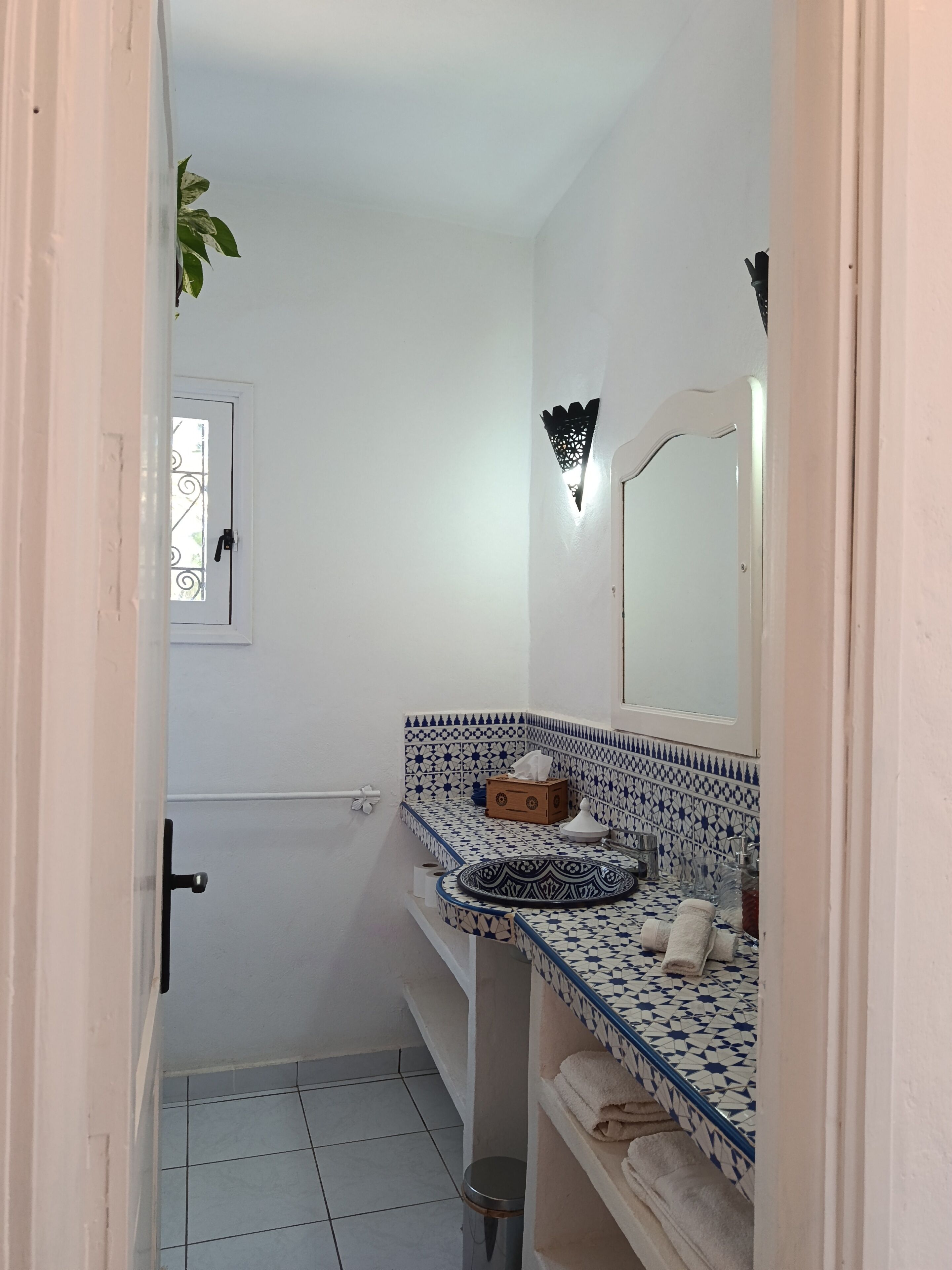 A bathroom with a white door, a window with a metal grill, a sink with blue and white patterned tiles, a mirror, and a small trash bin.