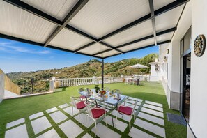 Eine überdachte Terrasse im Freien mit einem Tisch für zwei Personen, umgeben von Stühlen und Blick auf eine hügelige Landschaft.