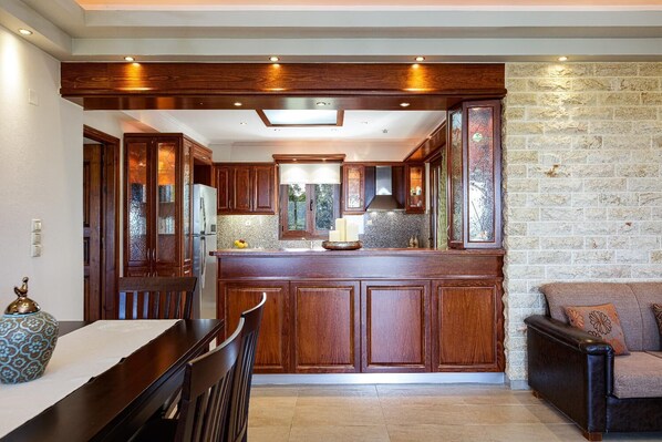 A kitchen with wooden cabinets, a stone wall, and a dining area with a table and chairs. - Villa Nefeli Breeze - Three Bedroom Villa, Sleeps 6 (Rethymno)