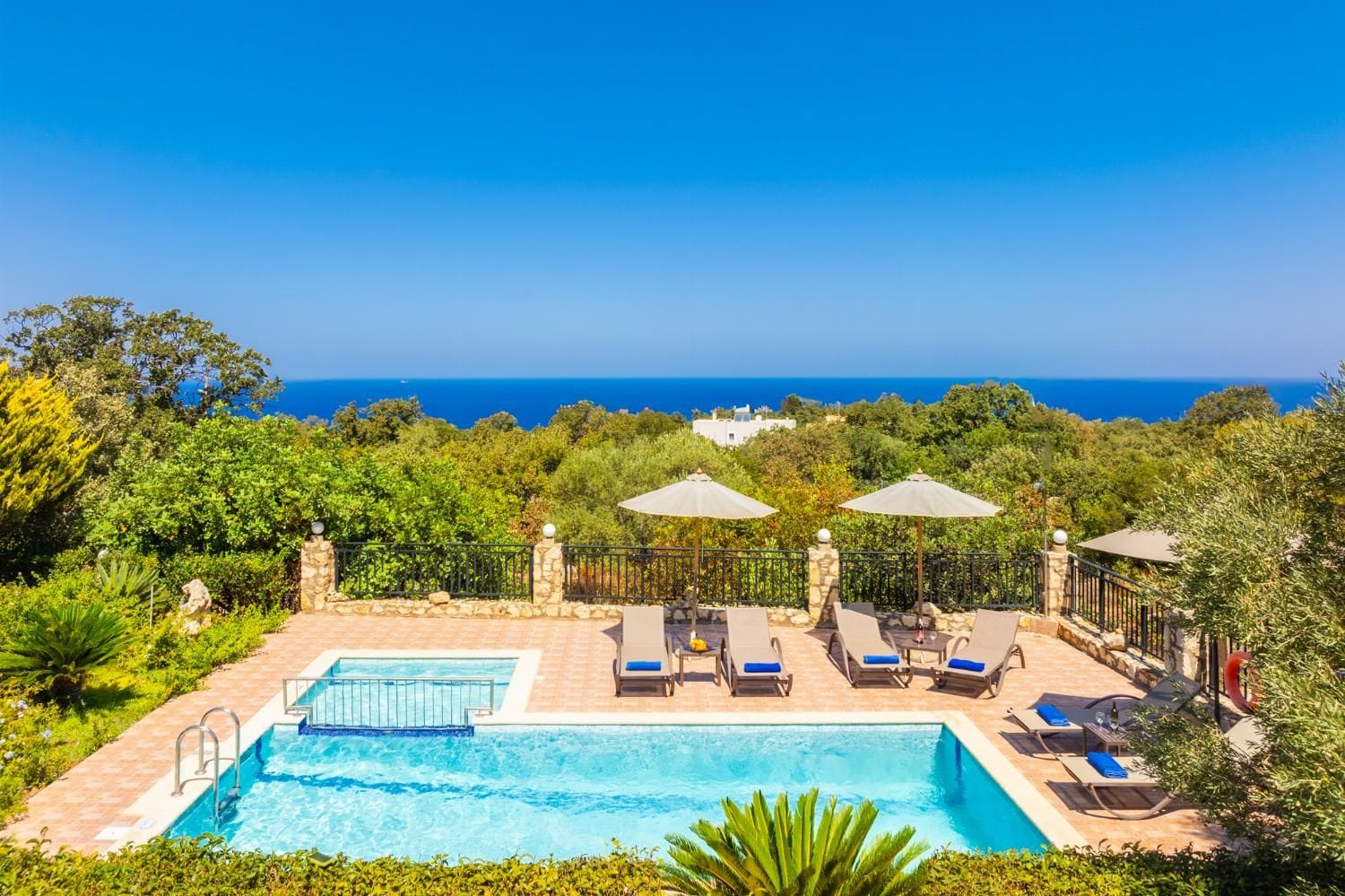 A swimming pool with lounge chairs and umbrellas, surrounded by greenery and overlooking a clear blue sea.
