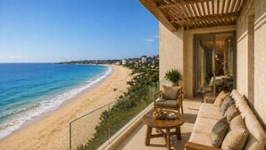 A balcony with a view of a beach and ocean, a sofa, a table with a bowl of fruit, and a chair.