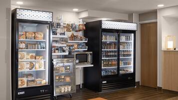 Een kitchenette met een magnetron, koelkast en automaten gevuld met snacks en drankjes.