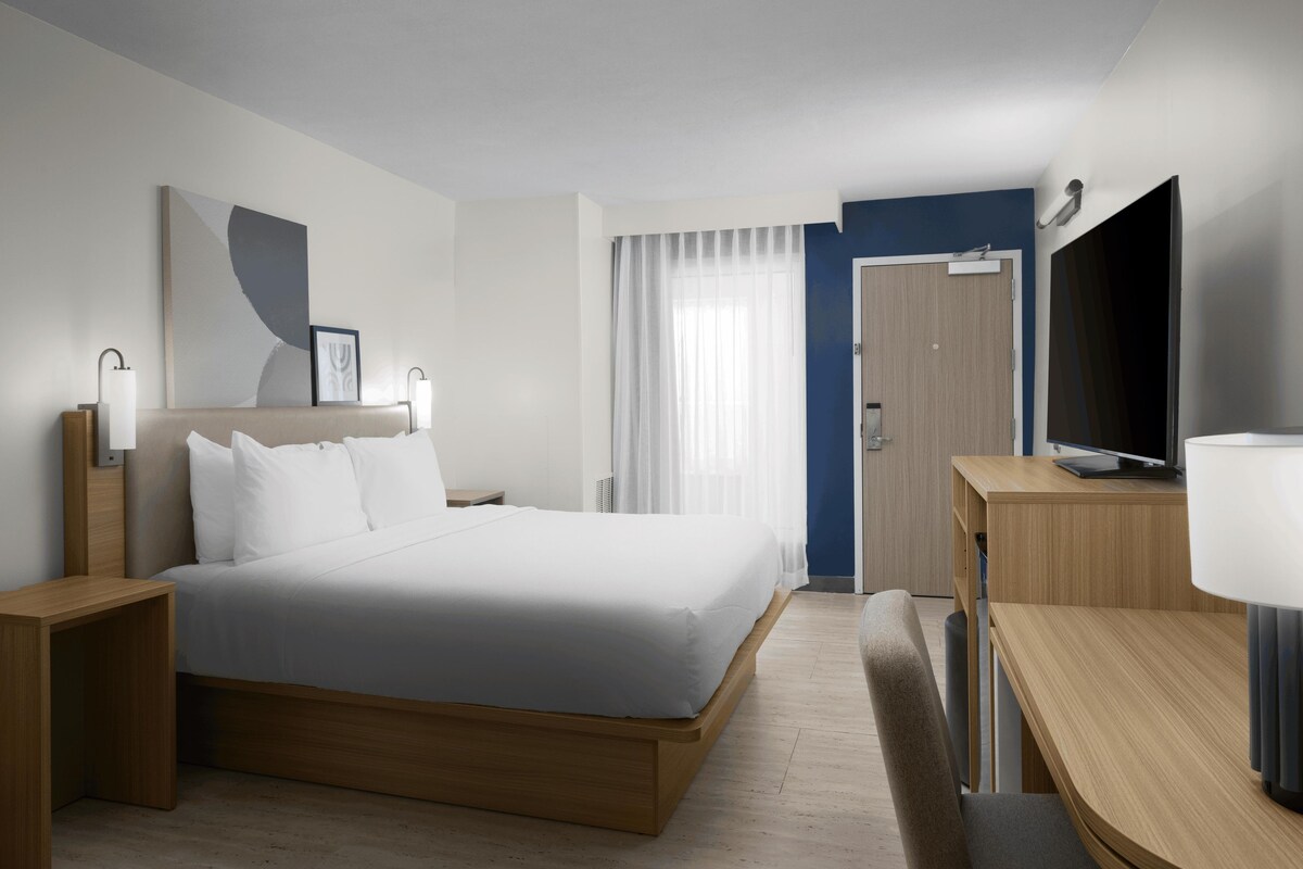 Habitación de hotel moderna con una cama grande, mesitas de noche, un escritorio con silla, televisor y una ventana con cortinas.