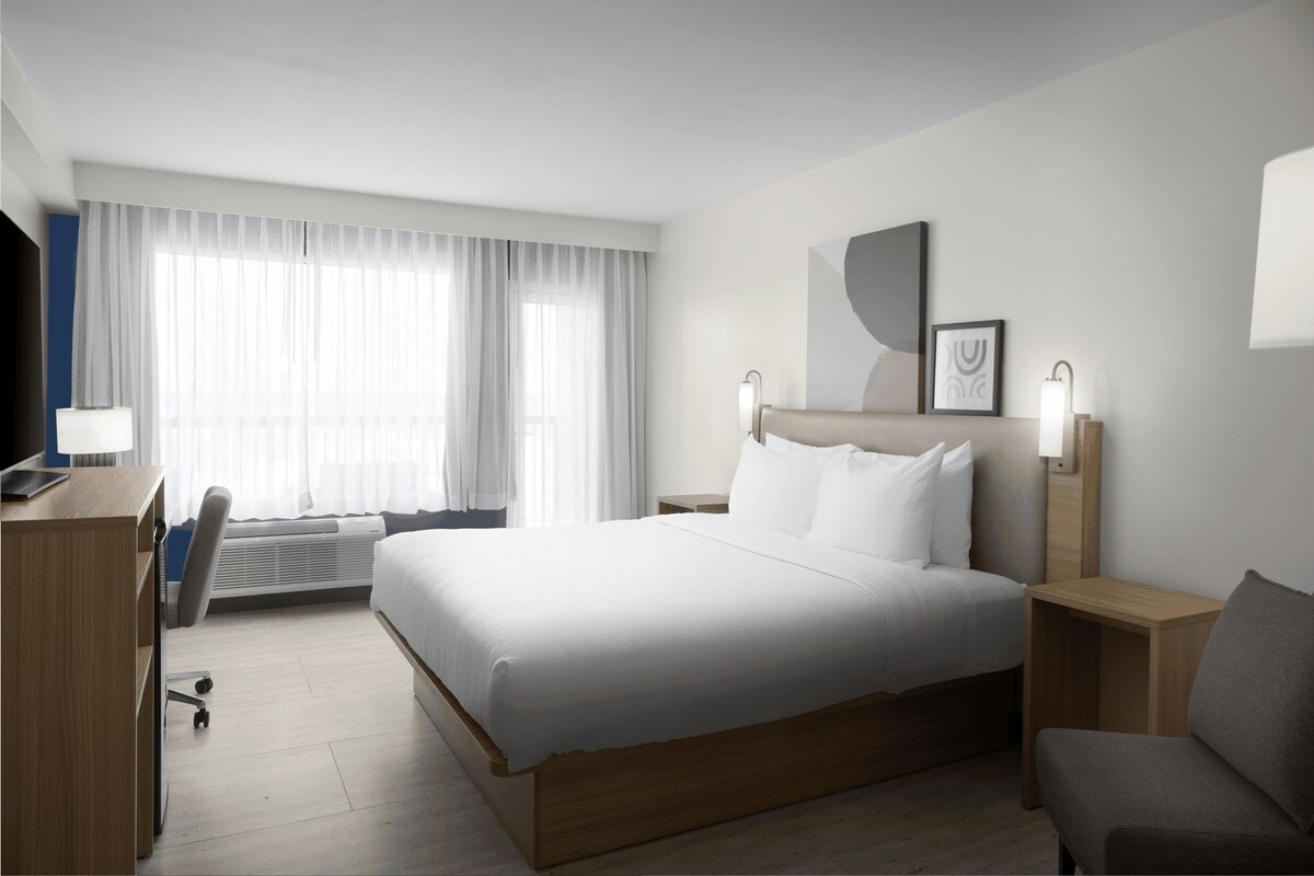 Habitación de hotel con una cama grande, un escritorio, una silla, un televisor de pantalla plana y una ventana con cortinas translúcidas.