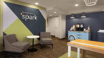A modern waiting area with a wall-mounted sign, two chairs, a small table, a counter with coffee dispensers, and a bicycle.