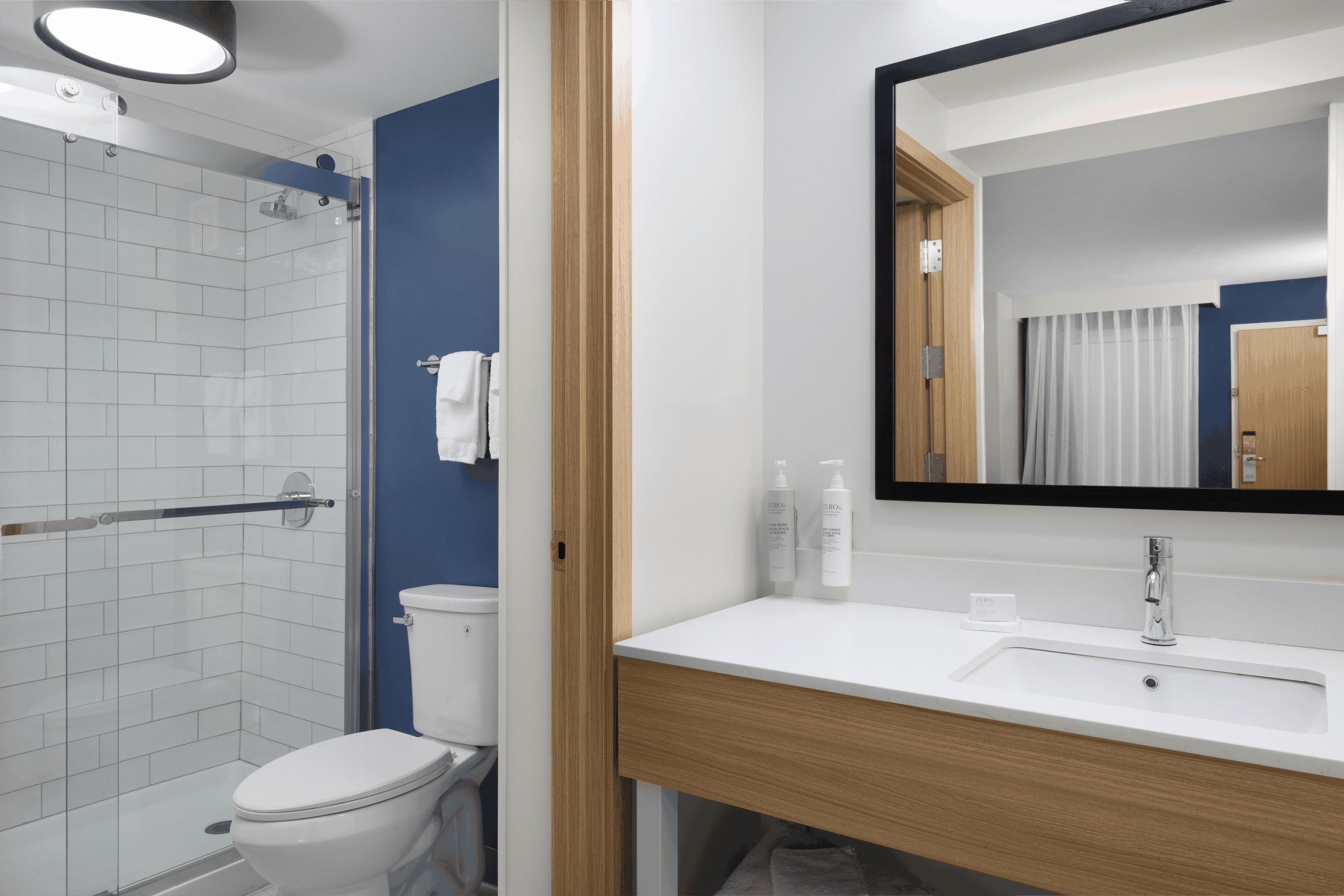 Un baño moderno con cabina de ducha de vidrio, inodoro blanco, tocador de madera con espejo y lavamanos blanco.