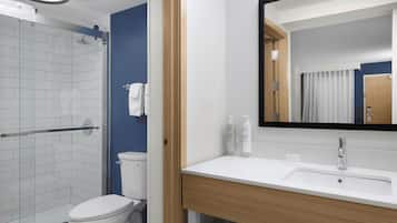 Un baño moderno con cabina de ducha de vidrio, inodoro blanco, tocador de madera con espejo y lavamanos blanco.