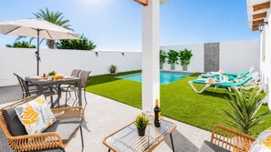 Eine Terrasse im Freien mit Esstisch, Stühlen und Sonnenschirm. Ein Swimmingpool mit Liegestühlen und ein kleiner Gartenbereich sind ebenfalls vorhanden.