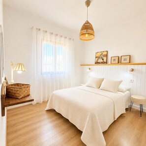 Ein Schlafzimmer mit einem Bett, einer Hängeleuchte, einem Nachttisch mit Lampe, einem Korbwäschekorb und einem Fenster mit Vorhängen.