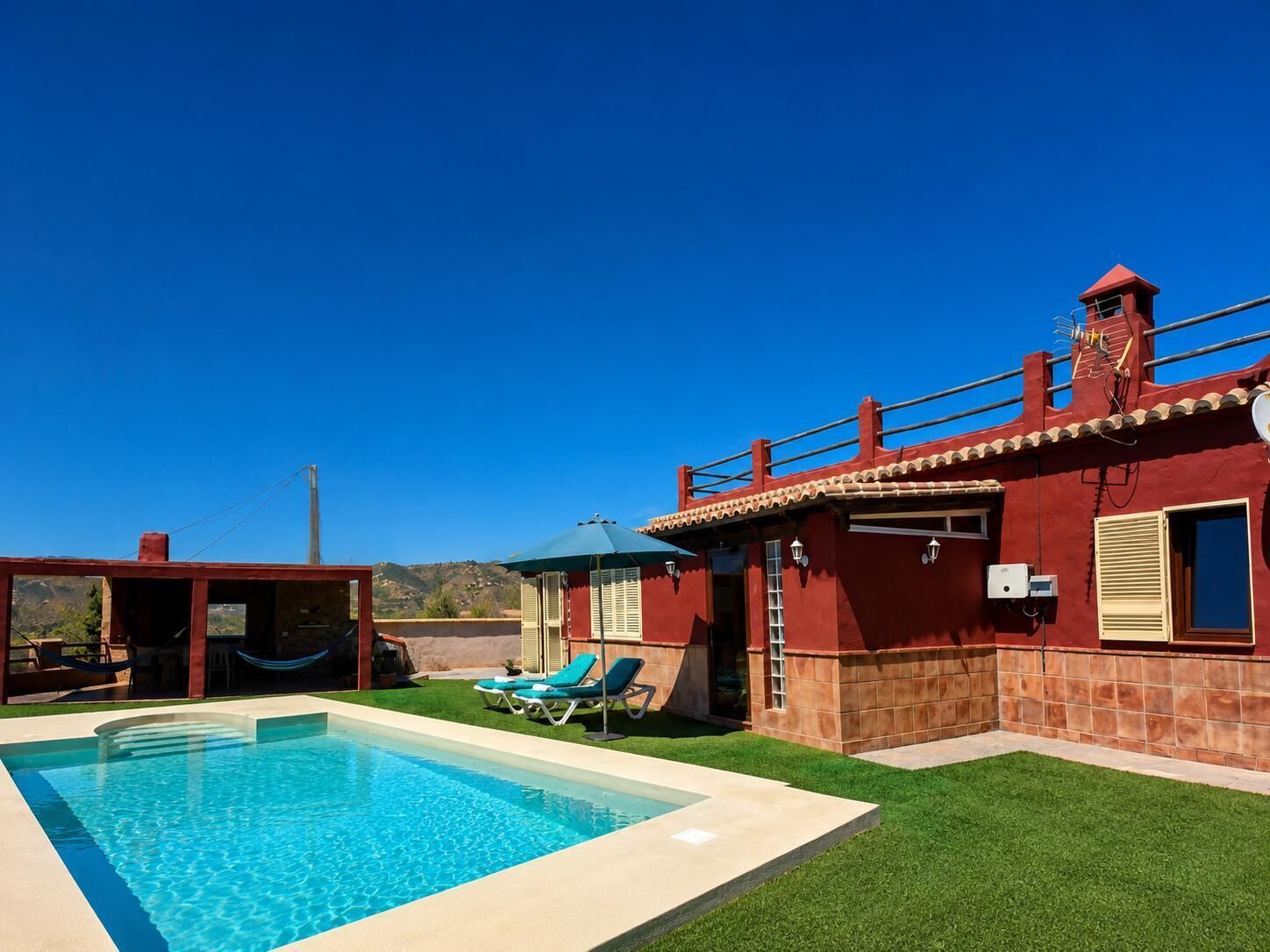 Una piscina con sillones de descanso, un edificio rojo con techo de tejas y un cielo azul despejado.