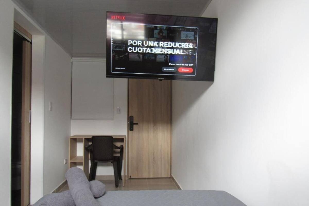 A room with a bed, a TV mounted on the wall displaying an advertisement, a desk with a chair, and a wooden door.