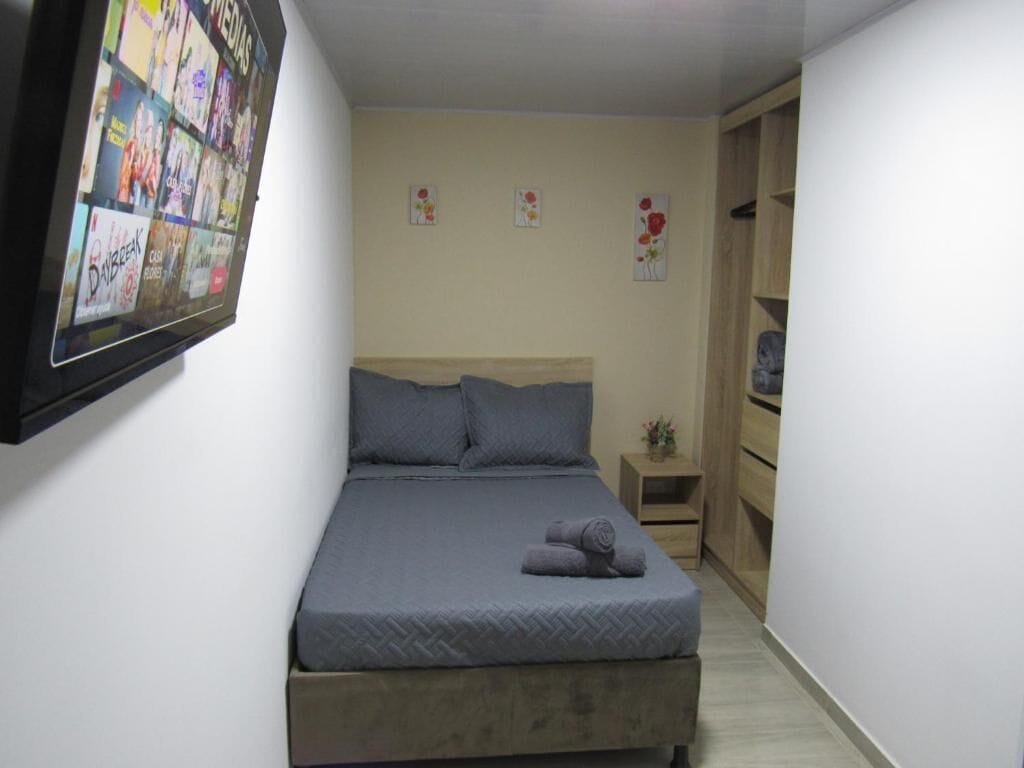 Um quarto com cama, uma mesinha de cabeceira, televisão na parede e um guarda-roupa de madeira.