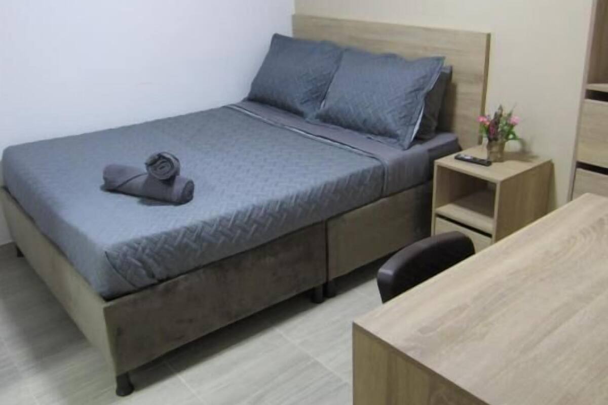 A single bed with a grey bedspread and pillows, a bedside table with a vase of flowers, and a wooden desk.