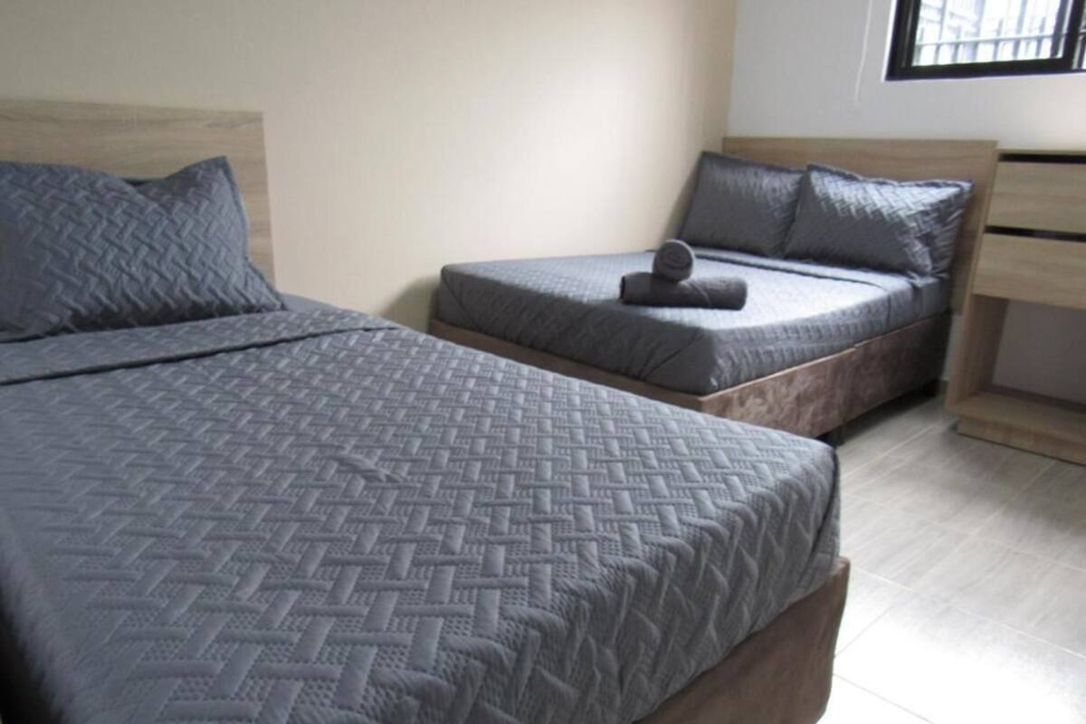 A double bed with a textured grey bedspread, a smaller bed with a similar grey bedspread, and a wooden nightstand with a drawer.