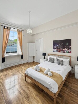 A hotel room with a large bed, a cityscape painting, a view of the city, a wardrobe, and a window with curtains.