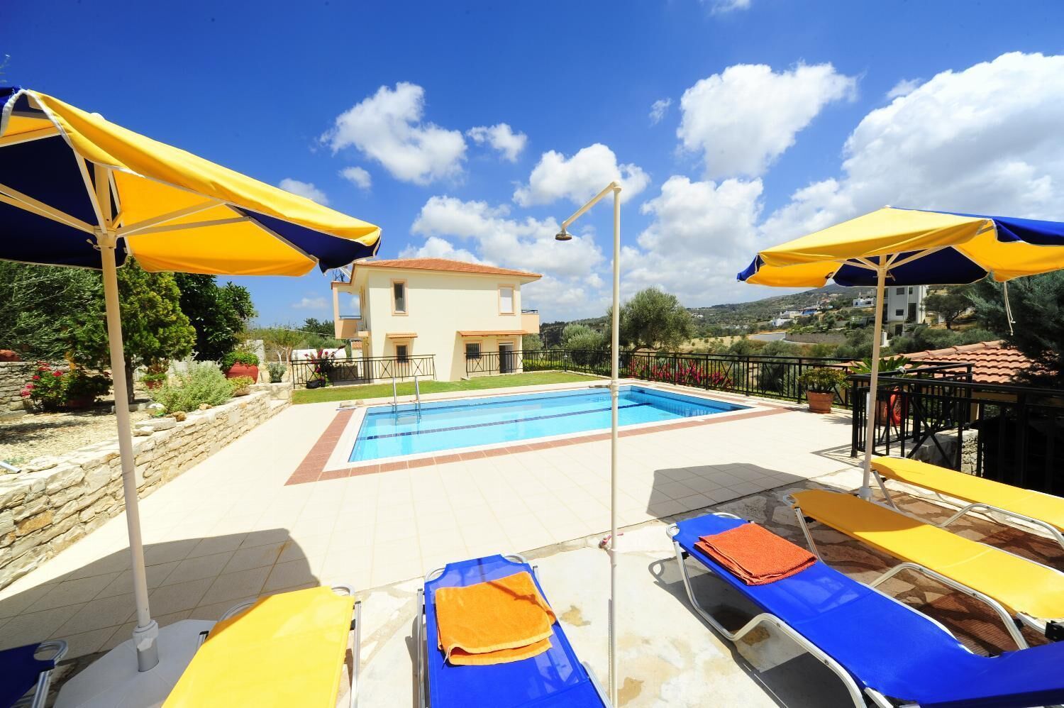 Area piscina con lettini e ombrelloni, una casa sullo sfondo e cielo sereno.