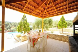 Eine überdachte Terrasse mit Esstisch, mit Blick auf einen Swimmingpool und eine Steinmauer.