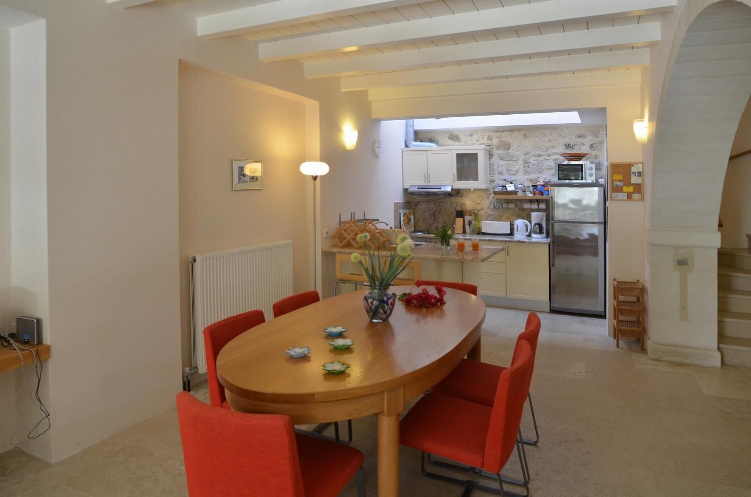 A dining area with a wooden table and red chairs, a kitchen in the background, and a stone wall.