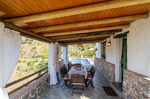 Um alpendre coberto com mesa e cadeiras, uma parede de pedra e vista para uma piscina e vegetação.