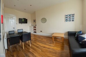 A living room with a wooden floor, a black sofa, a small wooden table, a TV mounted on the wall, and a dining area with a table and chairs. - Edinburgh Castle Views City Centre Apartment (Edinburgh)