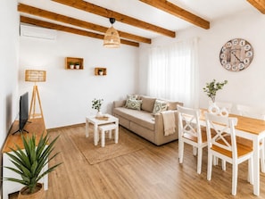 Ein Wohnzimmer mit Sofa, Couchtisch, Esstisch, Stühlen, Fernseher, Lampe, Uhr und einer Zimmerpflanze.