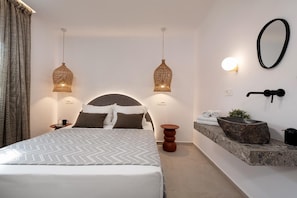 Ein modernes Hotelzimmer mit einem großen Bett, zwei Pendelleuchten, einem runden Spiegel, einem Steinsinkfeld und einer kleinen Topfpflanze.