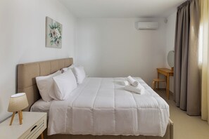 Ein ordentlich bezogenes Bett mit Kopfteil, ein Nachttisch mit Lampe, ein kleiner Schreibtisch mit Stuhl und eine Wand-Klimaanlage.