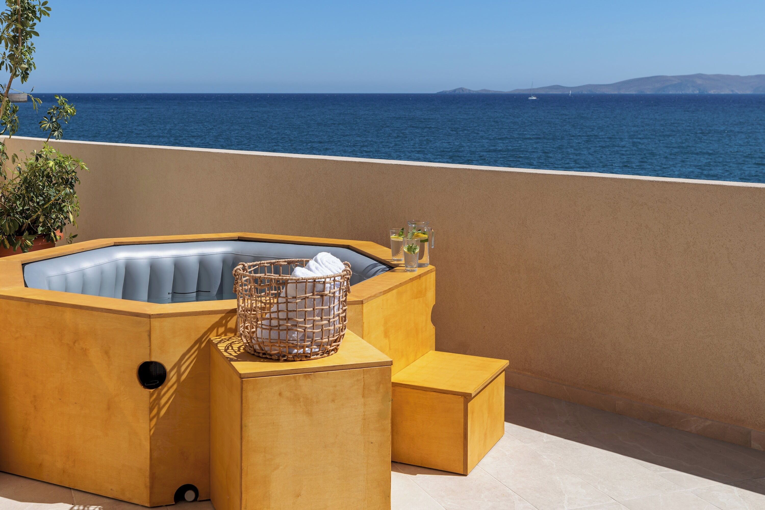 A hot tub with a wicker basket and a glass on a wooden platform, overlooking a body of water.