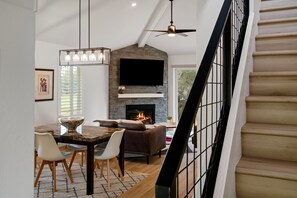 A modern living room with a fireplace, a dining area with a table and chairs, and a staircase with a railing.