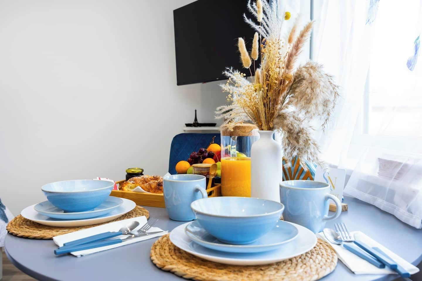 Desayuno con tazones, platos y tazas azules, una bandeja de frutas, una jarra de jugo de naranja y un florero con plantas secas.