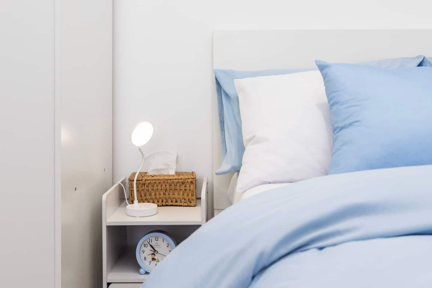 A neatly made bed with blue bedding, a white nightstand with a lamp, a clock, and a wicker basket.