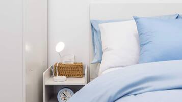 Cama bem arrumada com roupa de cama azul, mesa de cabeceira branca com uma luminária, um relógio e uma cesta de vime.