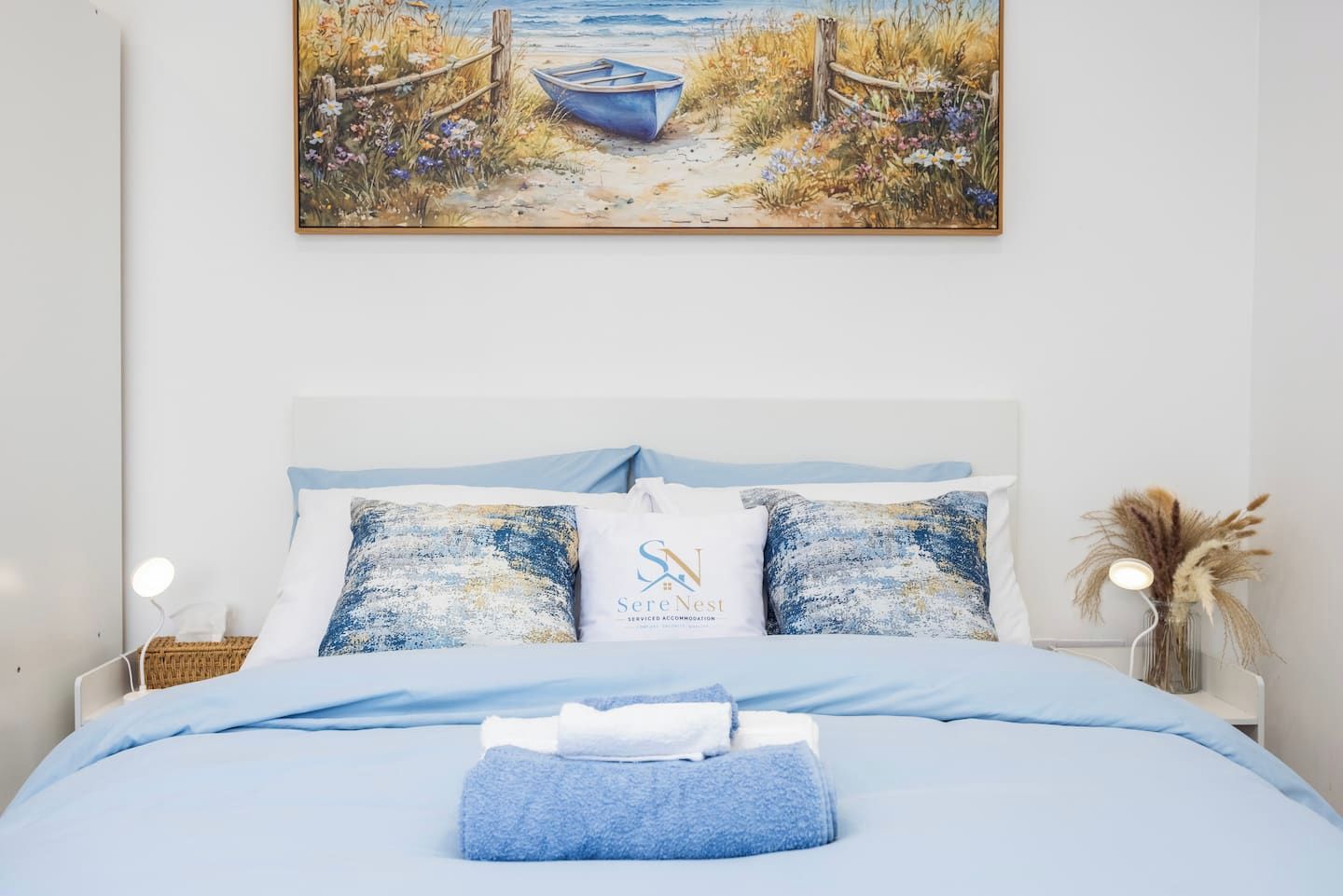Ein Schlafzimmer mit einem Bett, Kissen, einem Nachttisch, einer Lampe und einem Bild von einer Strandlandschaft.