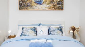 Un dormitorio con cama, almohadas, una mesita de noche, una lámpara y un cuadro con una playa.
