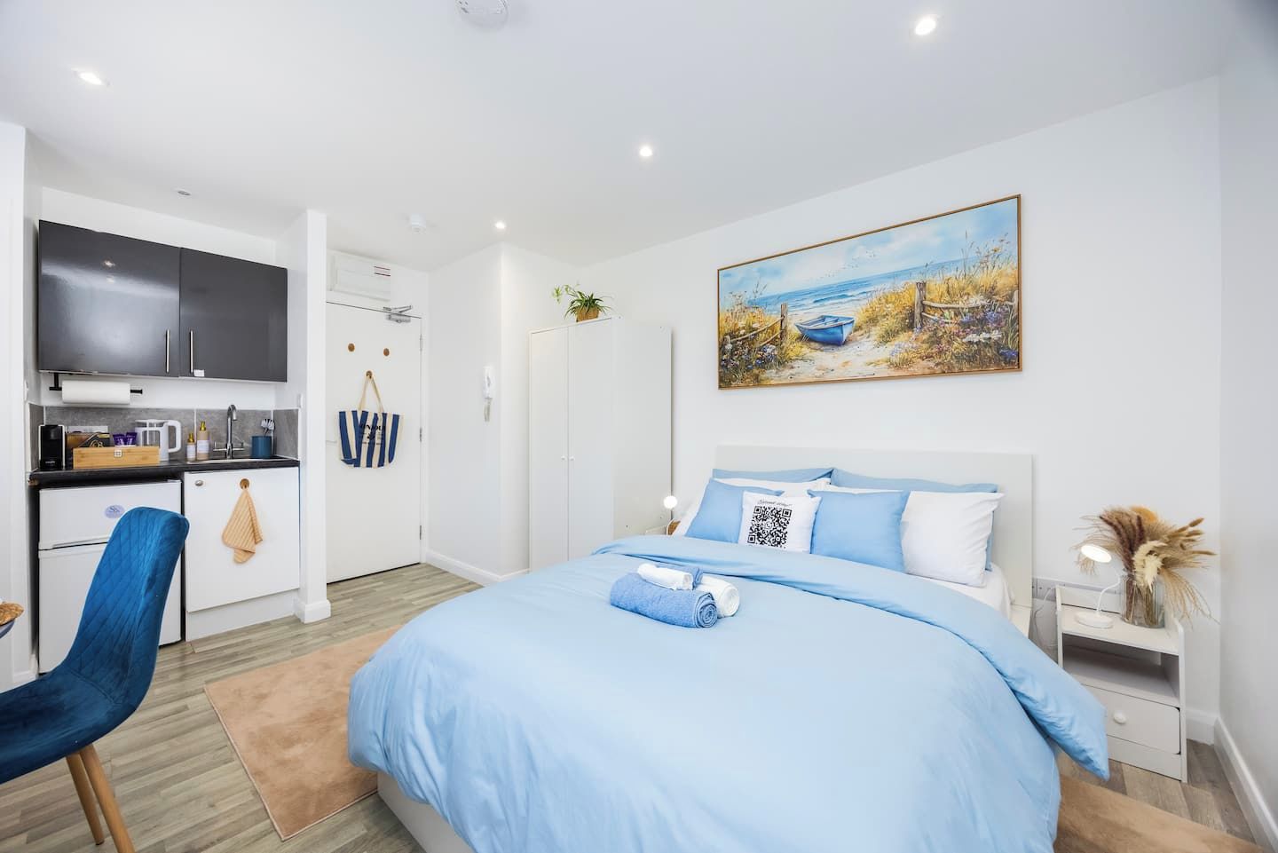 A modern bedroom with a large bed, a flat-screen TV, a small kitchenette, and a painting of a beach scene.