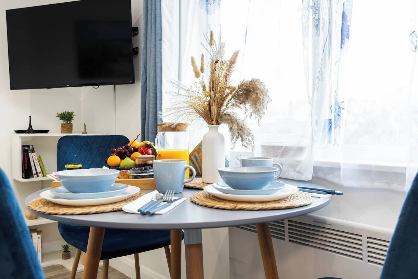 Uma área de refeições com mesa posta para o café da manhã, incluindo pratos, xícaras e uma cesta de frutas.