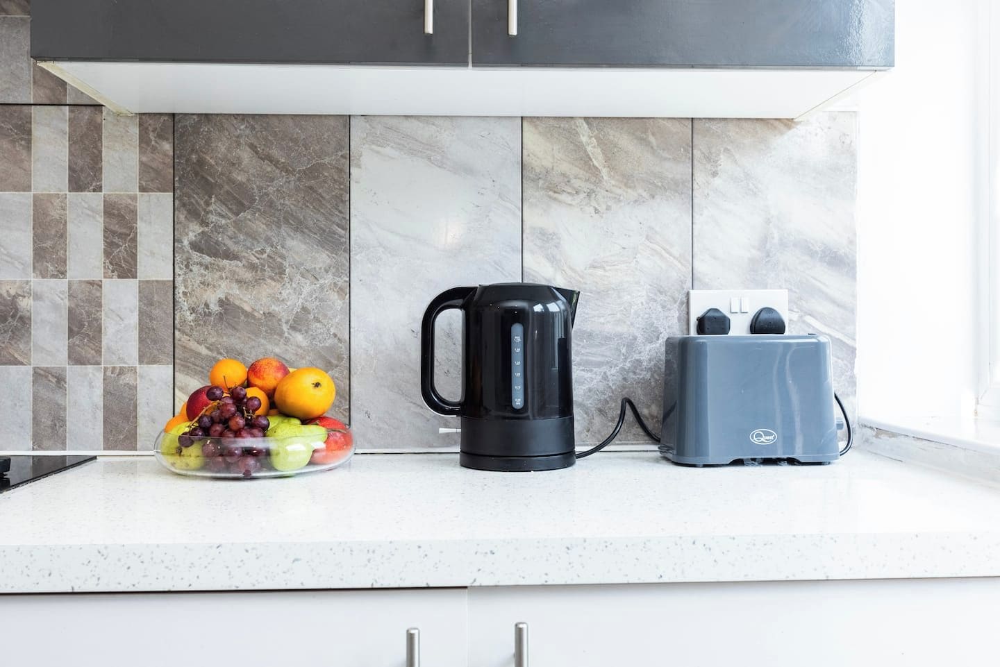 Una encimera de cocina con un frutero, una tetera eléctrica negra y un tostador.